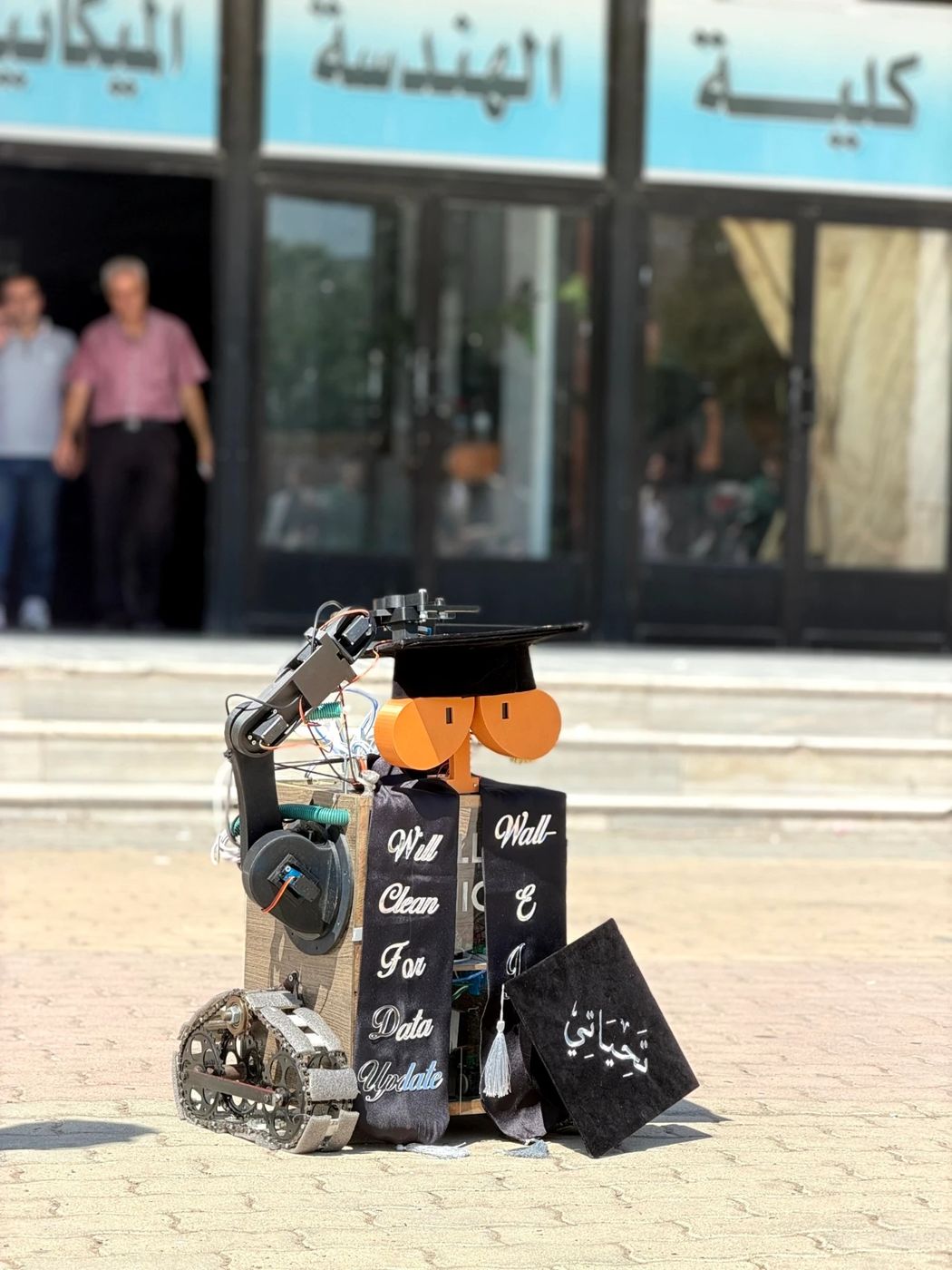 WALL-E Junior wearing a graduation cap and sash in front of the engineering faculty